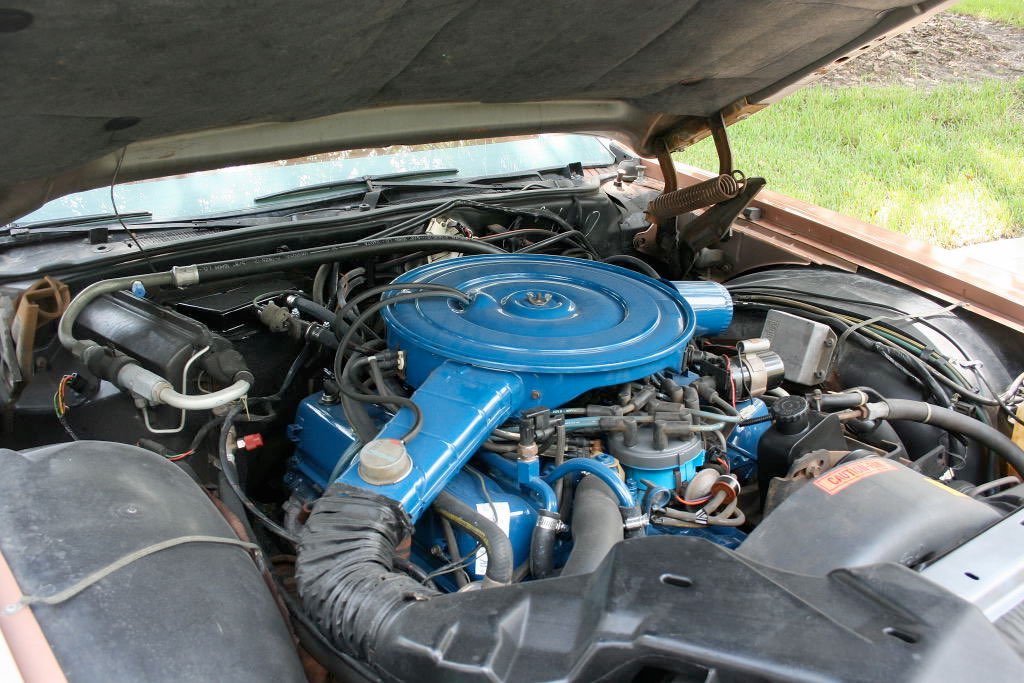 Under the open hood of a vintage car showing a blue engine with various hoses and metal components.
