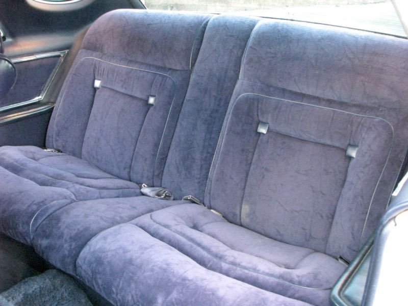 The backseat of a vintage car with plush, blue velvet upholstery and seat belts.