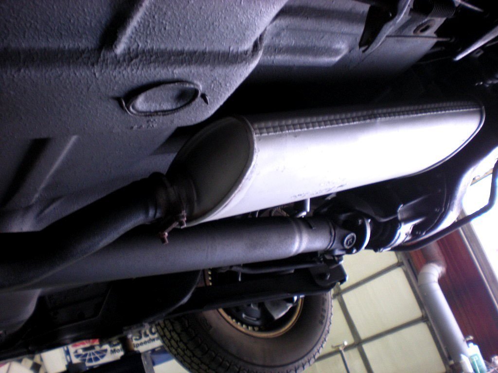 Under the vehicle, a close-up shows a silver muffler, black exhaust pipes, and part of a tire in the background.