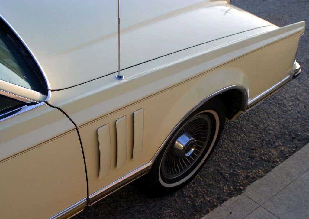 Close-up of the front left side of a vintage cream-colored car, showing the wheel, part of the hood, and side vents.