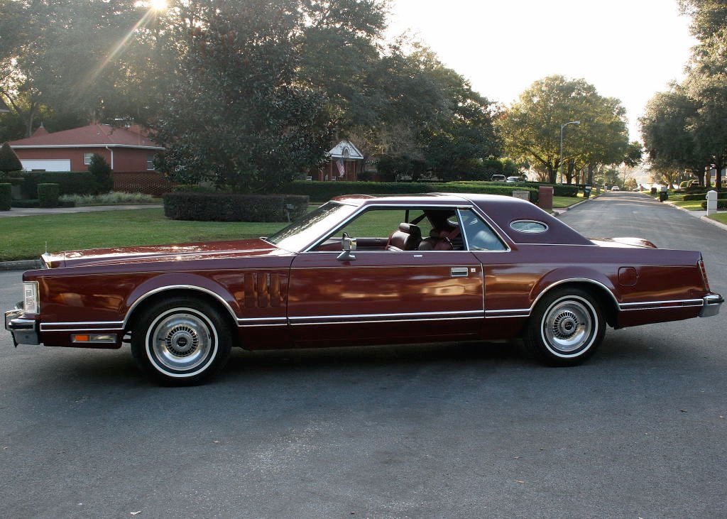 A vintage maroon-colored luxury coupe parked on a suburban street with trees and houses in the background, sunlight streaming through the trees.