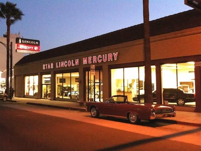 Exterior of a car dealership named Star Lincoln Mercury at dusk, with a vintage convertible parked in front and other cars visible inside through large glass windows.