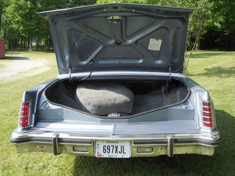 Rear view of a vintage silver car with the trunk open, showing a spare tire inside. The car has Ohio license plate 697XJL and a small AAA sticker on the bumper. The background includes grass and trees.