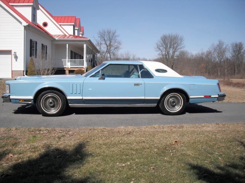 Light blue vintage convertible car with white roof parked on a paved driveway in front of a white house with a red roof and porch, trees in the background, clear sky.