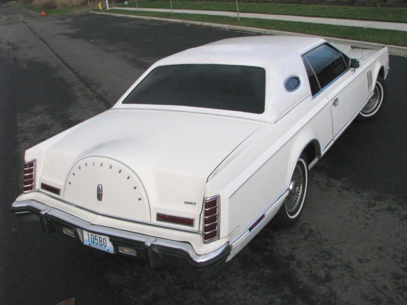 A white vintage car parked on a street, viewed from the rear and side angle, featuring a unique design on the back resembling a clock face or dashboard gauges.
