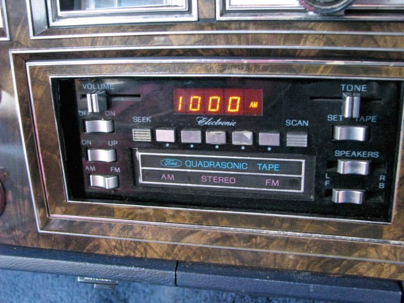 Vintage Ford electronic car stereo receiver with a woodgrain panel, displaying the radio station 1000 AM.