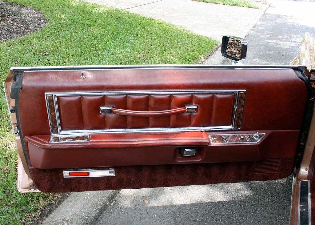 The interior door panel of a vintage car with brown faux leather, wood trim, and chrome accents, located next to a grassy area and sidewalk.