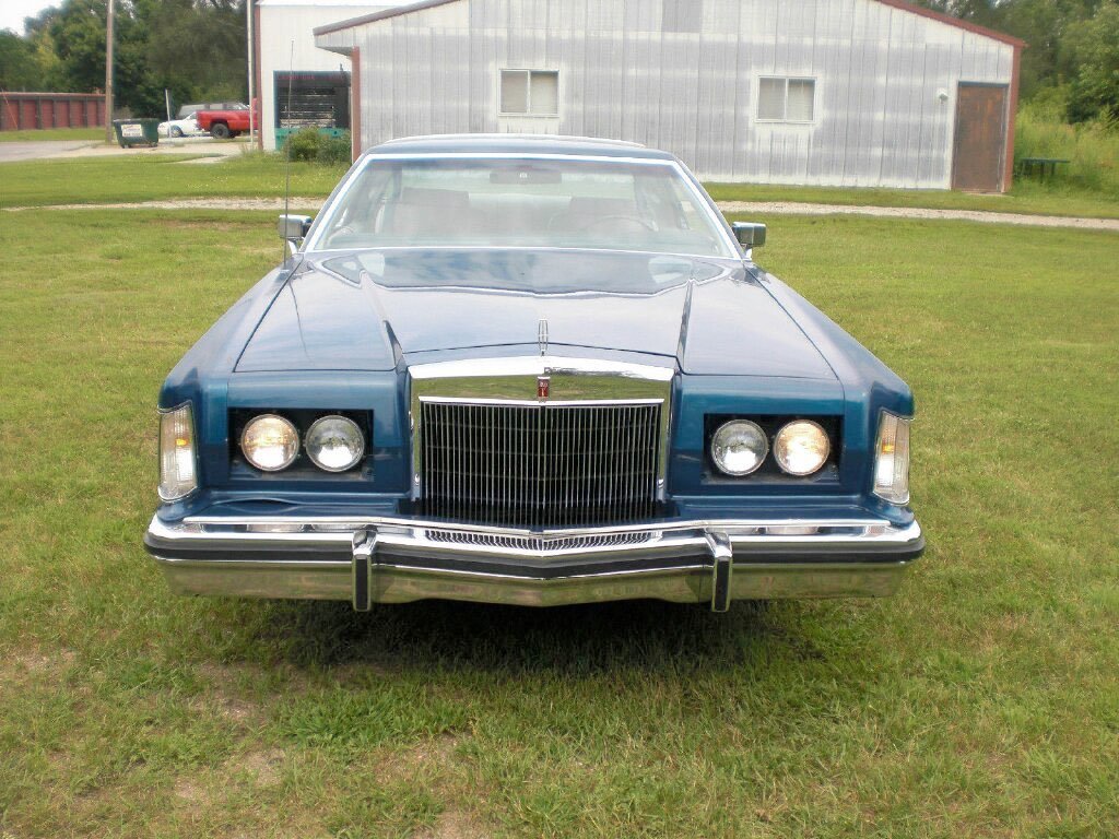 Front view of a vintage blue car with quad headlights and chrome grille parked on grass with a barn and trees in the background.