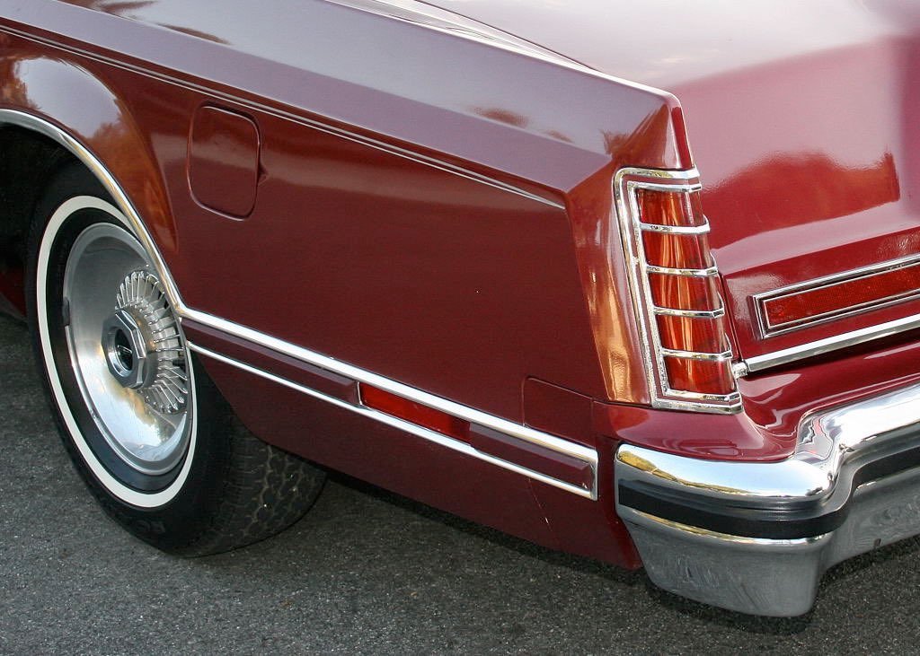 Close-up of the rear corner of a vintage red car, showing the wheel, tail light, and chrome bumper.