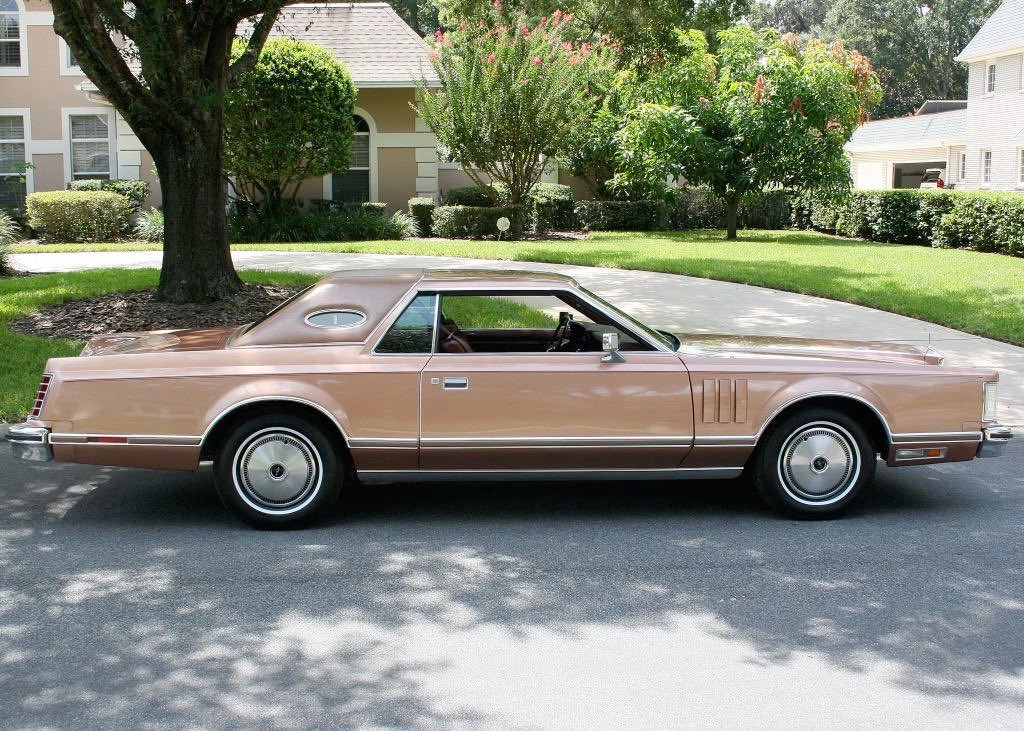 A vintage tan two-door car parked on a suburban street with green grass and trees in the background.