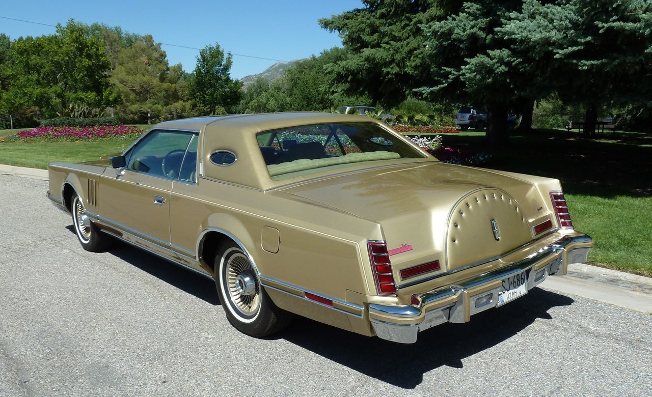 A vintage gold-colored Lincoln Continental Mark V car parked on a street with green trees and mountains in the background.