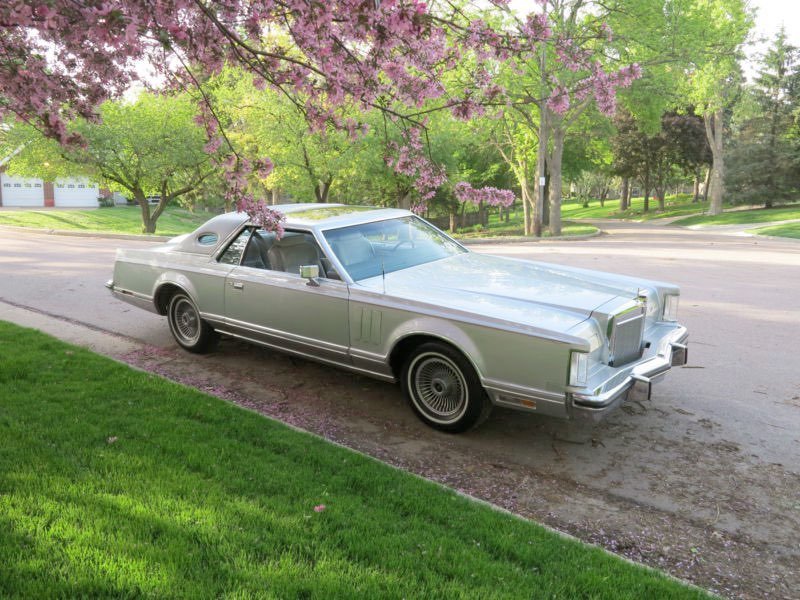 A silver vintage car parked on the side of a suburban street, with pink flowering trees and green lawns in the background.