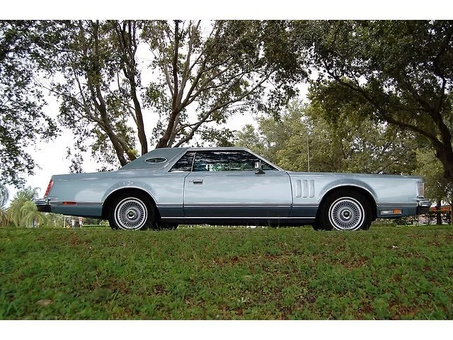 A vintage light blue car parked on grass under trees with green foliage.