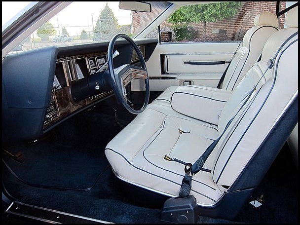 Interior of a vintage car with white leather seats, black steering wheel, and dashboard with wood trim.