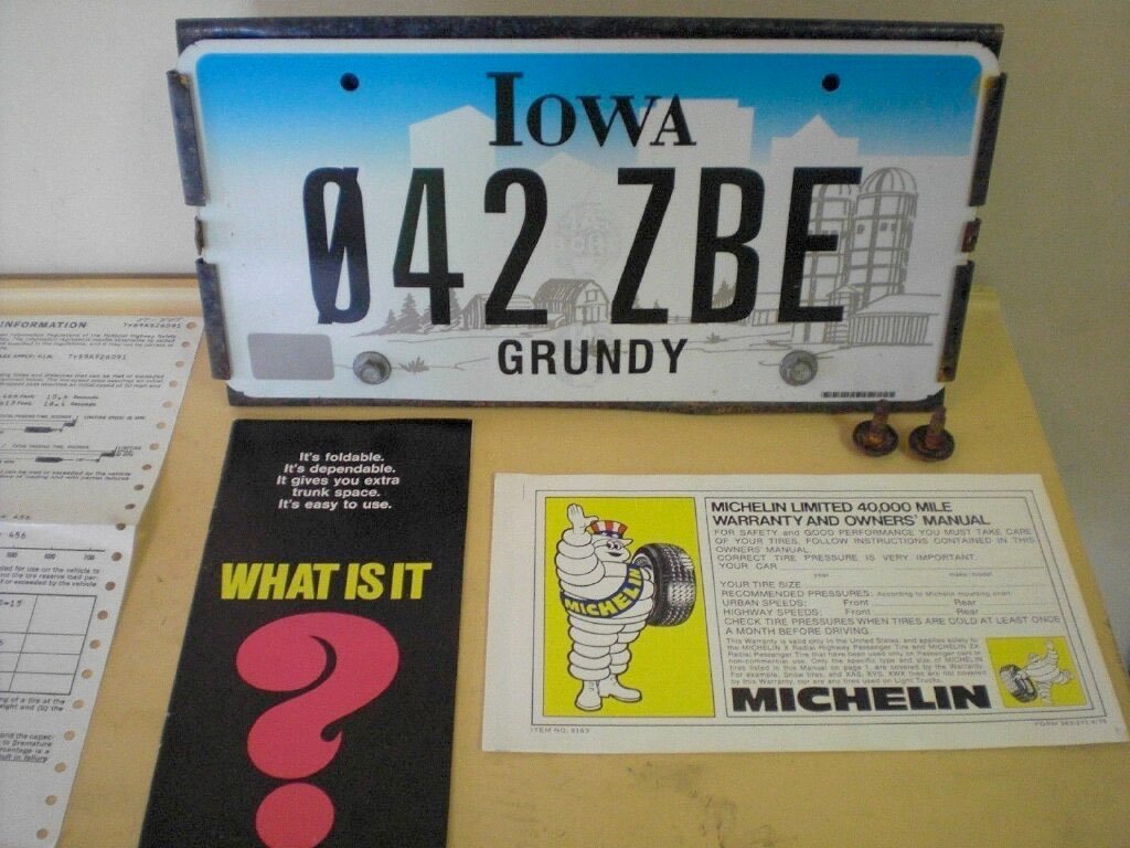 An Iowa license plate with the number 042 ZBE and a Grundy county label, a black pamphlet with a pink question mark asking "What is it?" and a yellow and white Michelin tire warranty and owner's manual on a table.