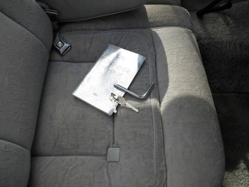 A gray car seat with a metal travel mug, a peephole lock, and a metal L-shaped tool on it. The tool is holding a key with a white paper tag. A book or notebook labeled "Optional Travel" is also on the seat.