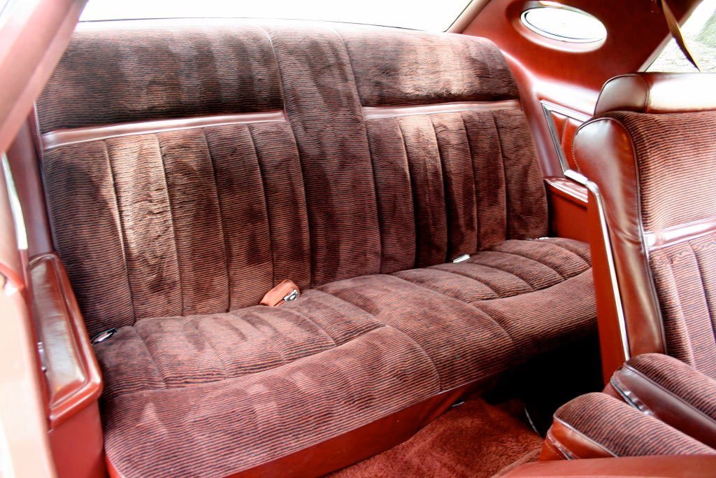 The back seat of a vintage car with brown velvety upholstered bench seating and matching interior details.
