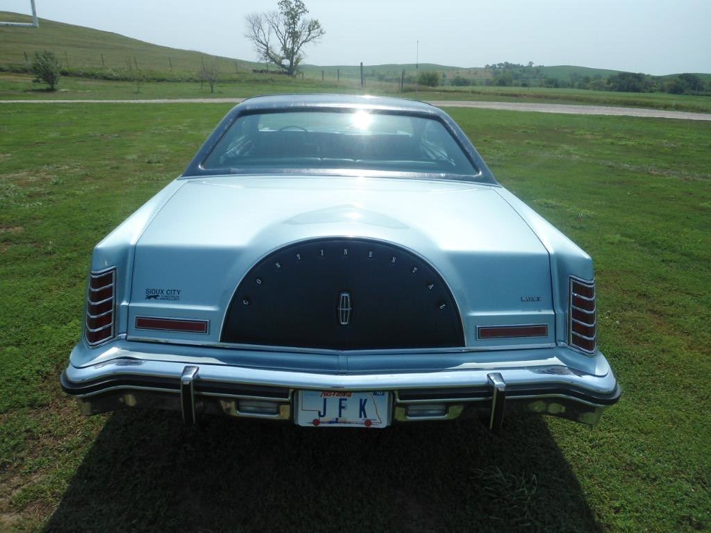 Rear view of a vintage light blue Lincoln Mark VI car with a spare tire cover, parked on grass with a rural landscape in the background.