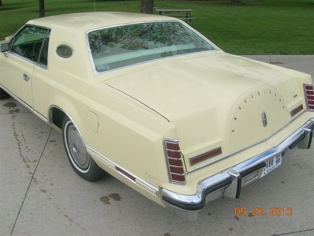 A vintage yellow car parked on a sidewalk in a park with grass and trees in the background.