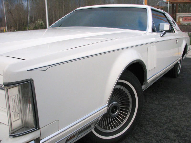 A classic white luxury car with wire-spoked wheels and a rectangular headlight design, parked outdoors with a fence and trees in the background.