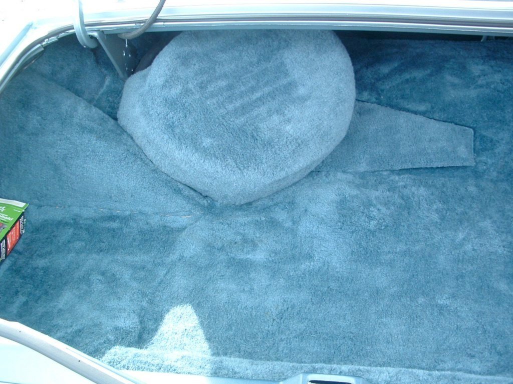 Inside the trunk of a car with blue carpeting, a large round blue pillow, and a small portion of a green box on the left side.