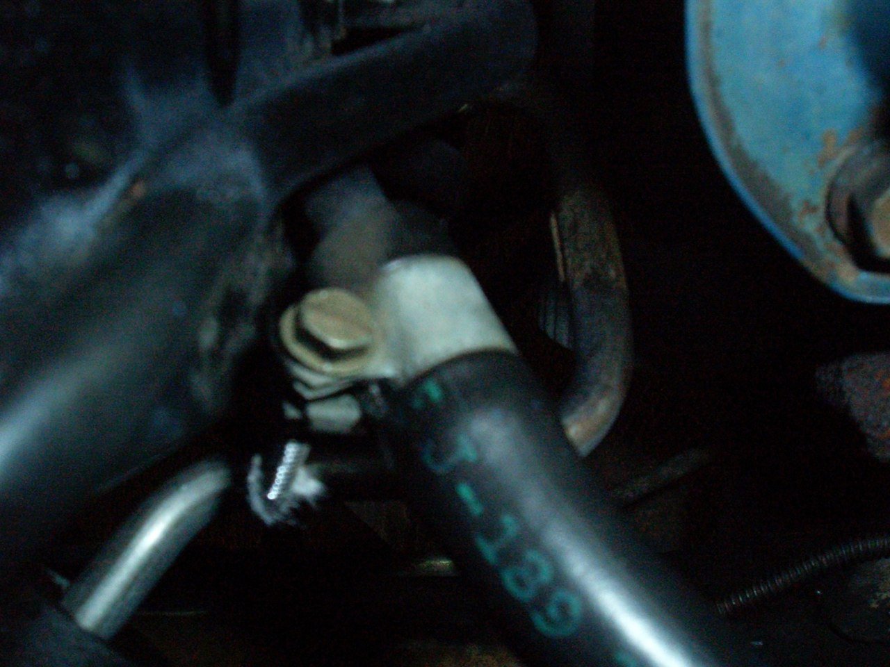 Close-up of a vehicle engine component, including a bolt, a black hose, and a metal tube with visible wear and rust.
