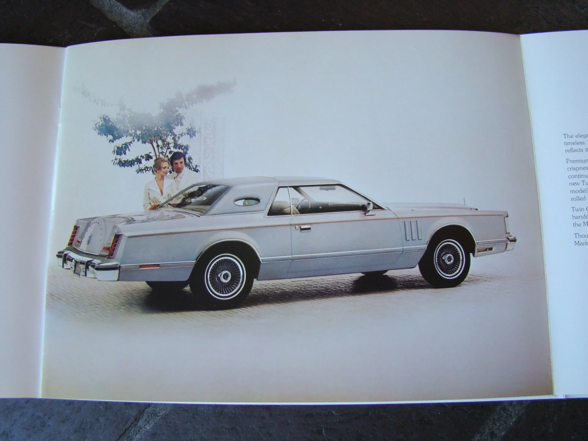 A vintage silver car with a partially closed roof, parked on a street with a person dressed in white standing beside it, and a tree in the background.