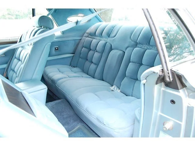 Vintage car interior with light blue upholstered backseat and side panels, with visible seatbelt and car window.