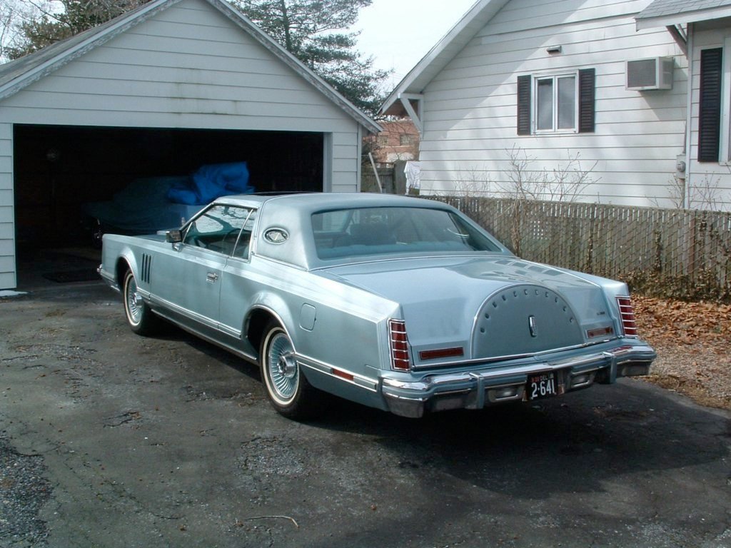A vintage silver coupe car parked in a driveway in front of a garage and house.