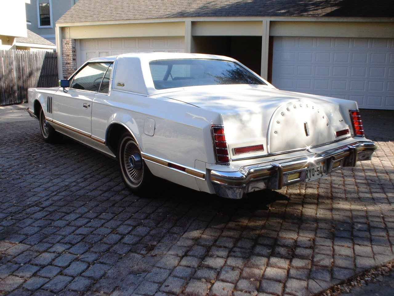 A vintage white Lincoln Mark V coupe parked on a cobblestone driveway in front of a house with a white garage door.