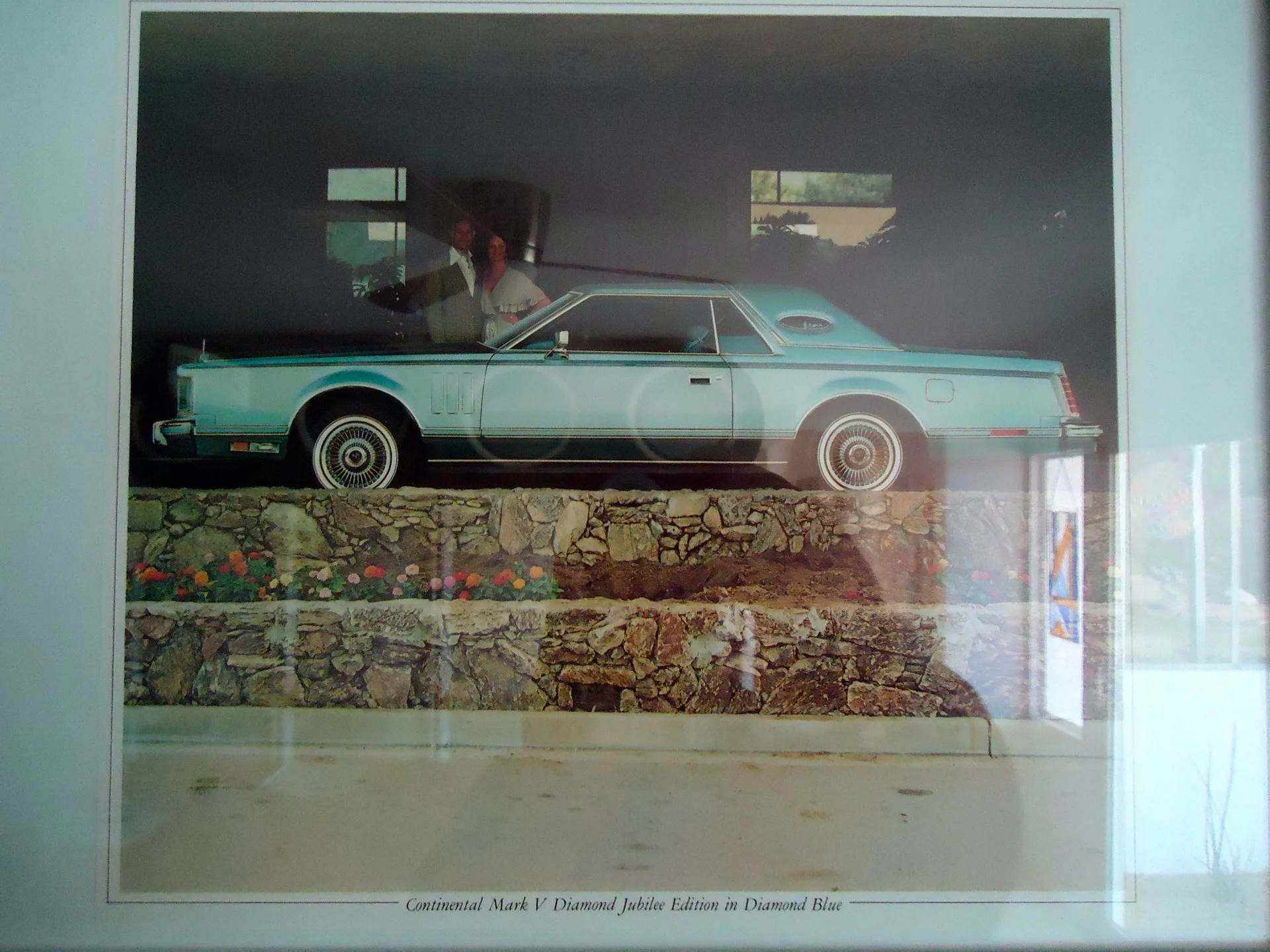 A framed photograph of a vintage turquoise car, a Continental Mark V Diamond Jubilee Edition in Diamond Blue, displayed on a tiered stone platform with a flower bed underneath. Two people are reflected in the glass of the frame, standing behind the p