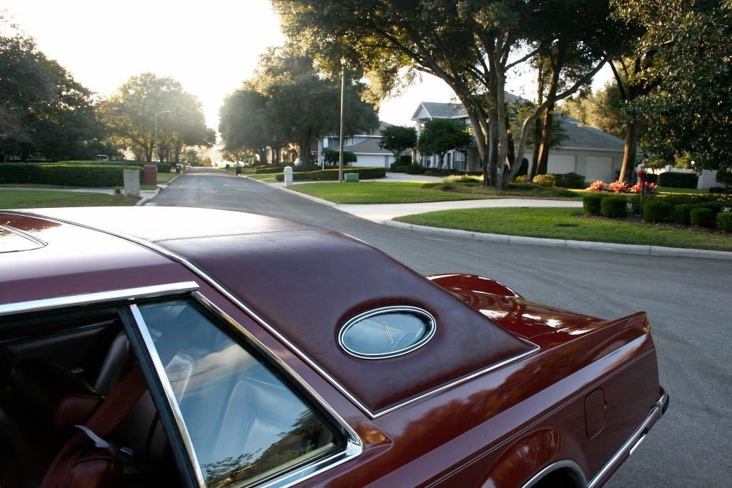 Close-up of the rear part of a vintage brown car parked on a suburban street with green lawns, trees, and houses under a clear sky.