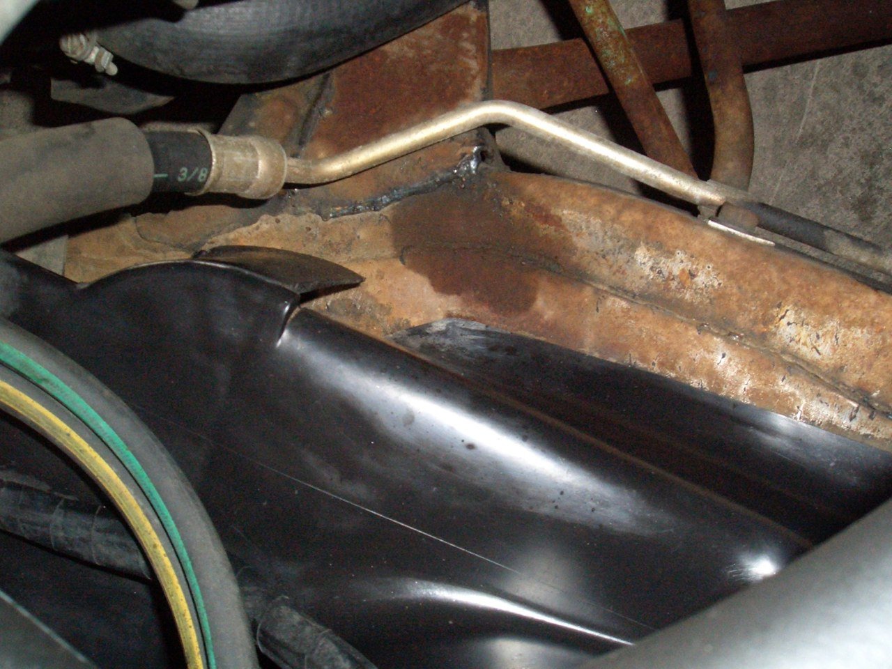 Close-up of a rusty metal surface in a vehicle, with a black plastic part, a silver tube, and a rubber hose.