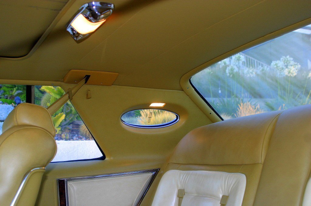 Interior of a vintage car with yellow leather seats, small oval window, rear window showing outdoor garden, and ceiling light.