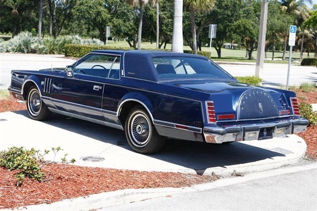 A vintage dark blue luxury coupe car parked on a street in a tropical park setting.