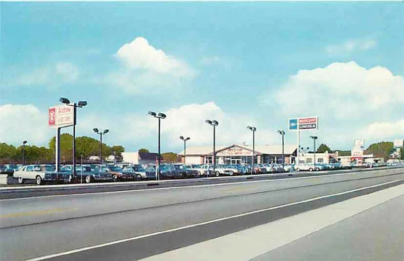 A car dealership lot filled with various cars parked in rows, with a large sign and a building in the background, along a highway under a partly cloudy sky.