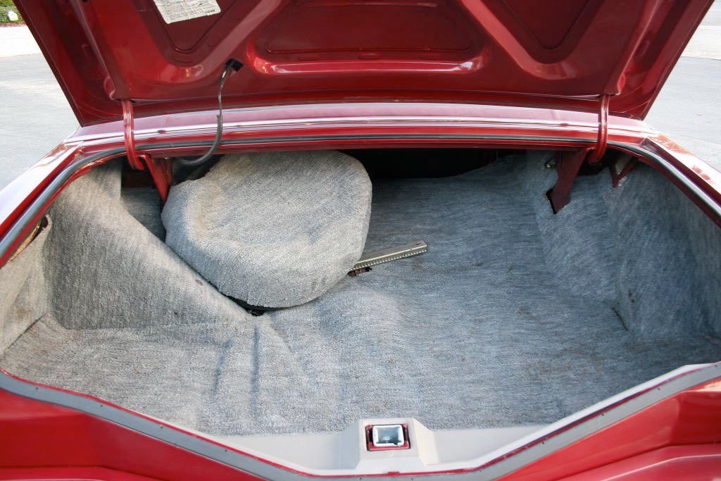 Open car trunk showing a spare tire covered with gray fabric and a metal tool inside the trunk, all lined with gray carpeting.