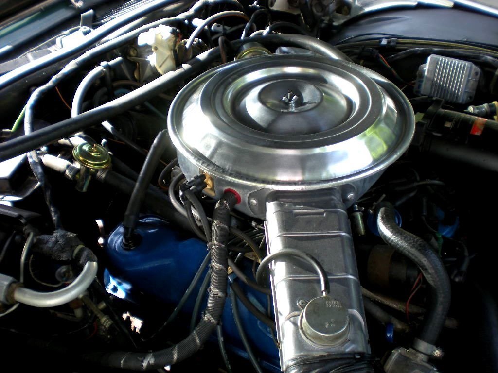 Close-up view of a car engine with a round, silver air cleaner on top, various hoses, wires, and metallic components surrounding it.