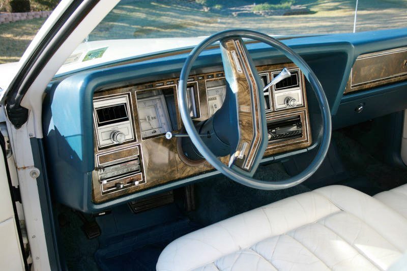 Interior of a vintage car with a woodgrain dashboard, steering wheel, and white leather seats.