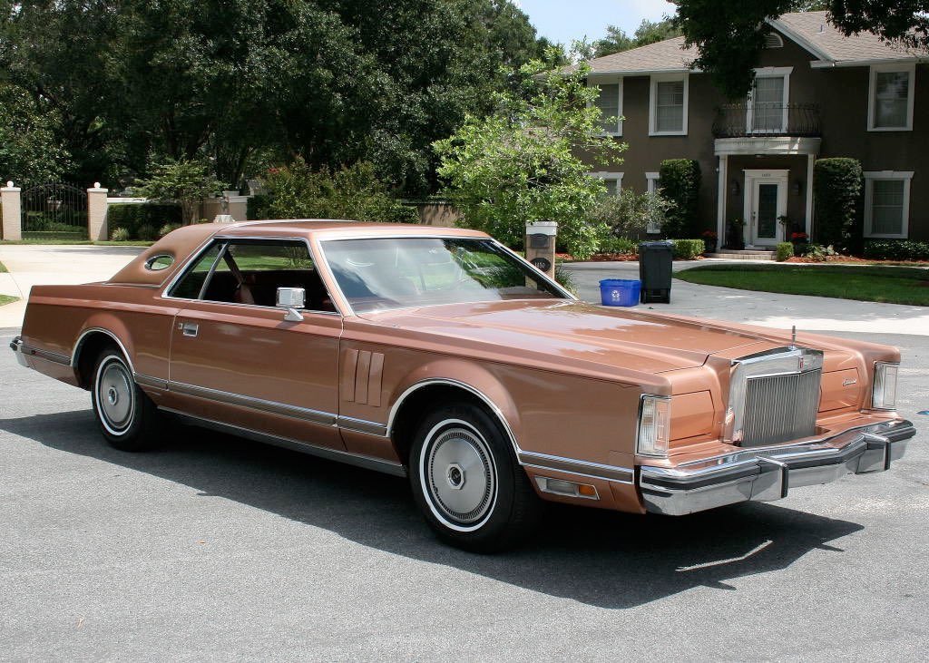 A vintage brown 1970s luxury sedan parked on a suburban street with a house and trees in the background.