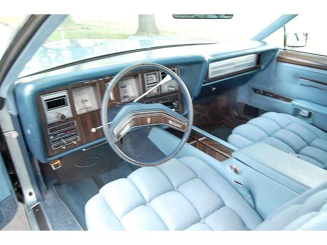 The interior of a vintage car featuring light blue plush seats, a wood grain dashboard, and steering wheel with analog gauges.