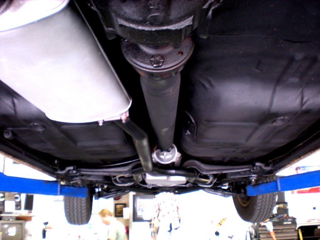 Underneath view of a car showing the exhaust system, drive shaft, and underside panels, with the car lifted on a hydraulic lift in a garage.