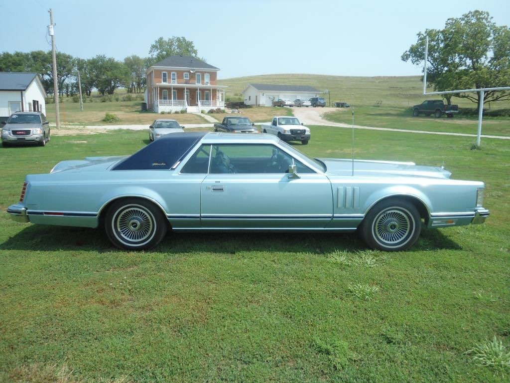 A vintage light blue two-door coupe car with black vinyl roof parked on green grass, with a two-story house, several other parked cars, and trees in the background.
