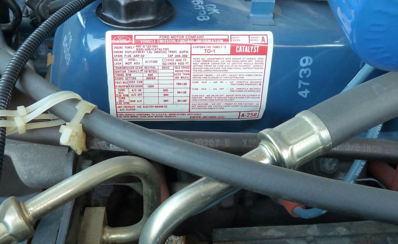 Close-up of a vehicle's engine compartment showing a Ford motor company emission control information sticker, various tubes, and metal components.