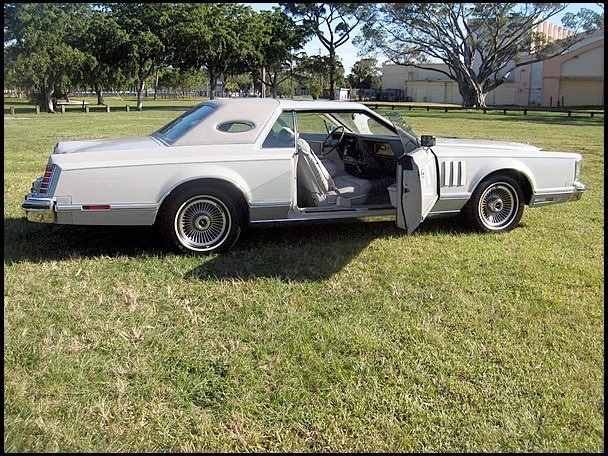 A vintage white luxury car with its front door open, parked on a grassy area with trees and buildings in the background.