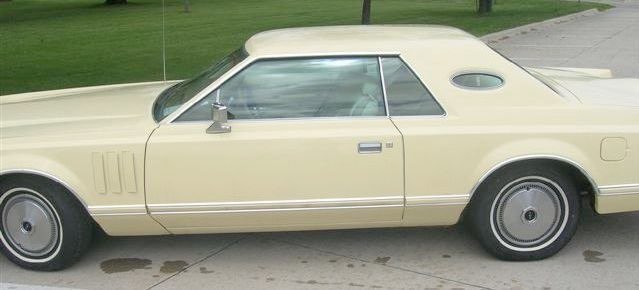 A vintage cream-colored two-door car with a hardtop, parked on a concrete surface near a grassy area.
