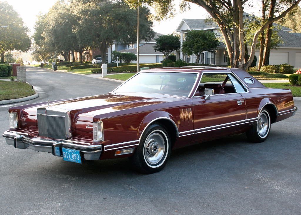 A vintage maroon luxury car, likely a 1970s Lincoln Continental, parked on a suburban street with houses and trees in the background.