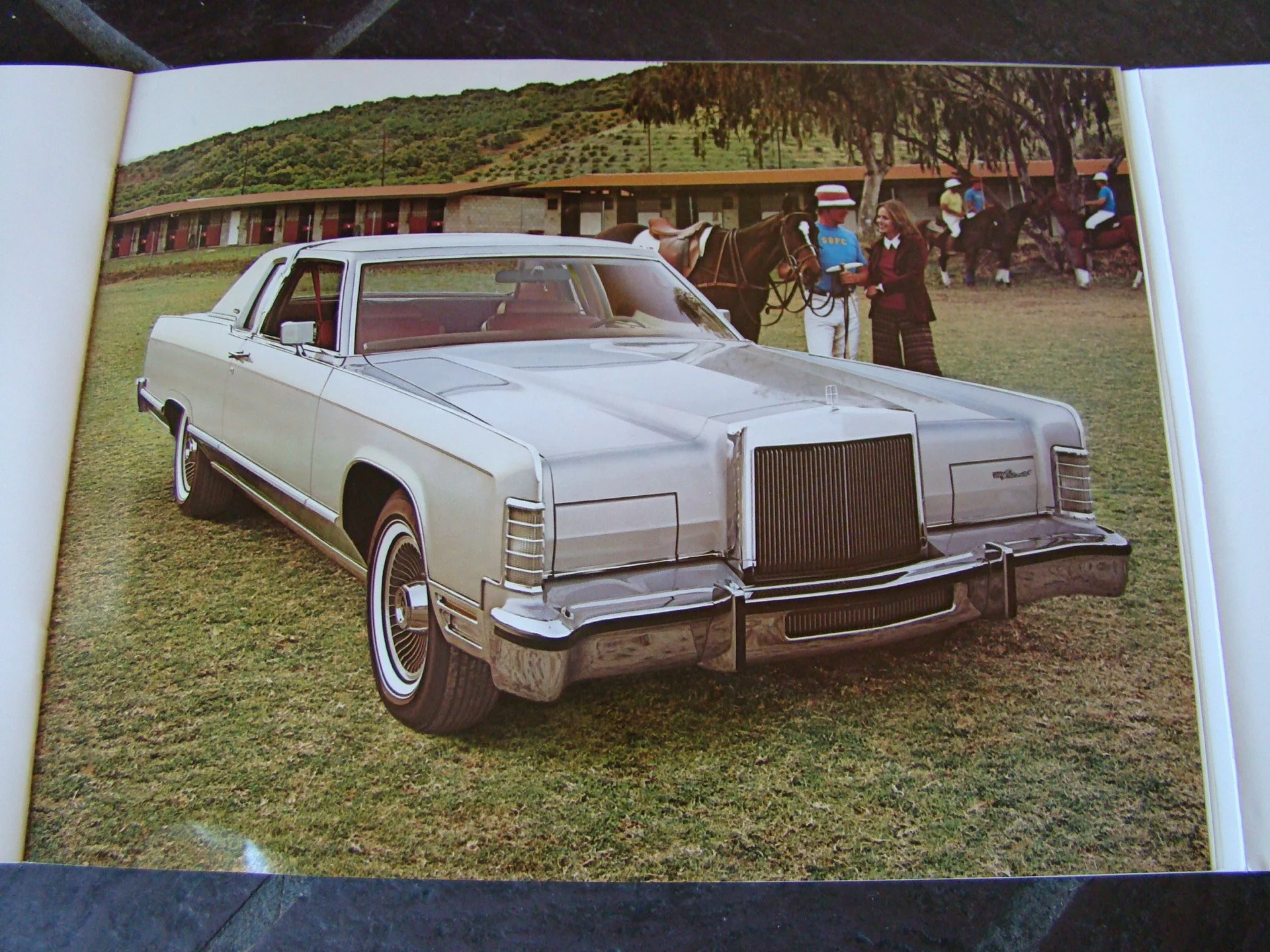 A vintage silver limousine parked on grass with a background of people, horses, and greenery.