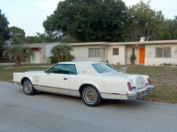 A white vintage car parked in front of a mid-century modern house with palm trees.
