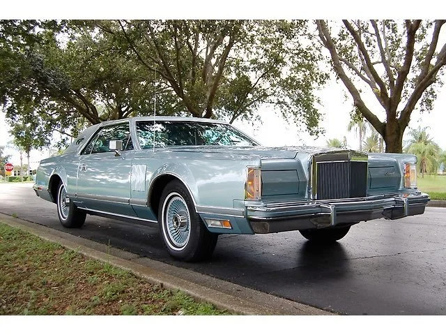 A vintage light blue luxury car parked on the side of a road under large trees.